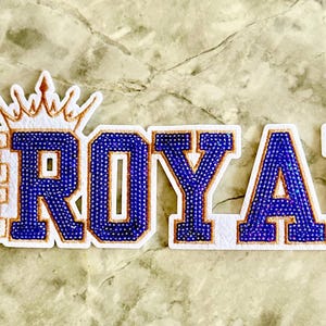 May include: White felt patch with the words "We Are Royal" in gold embroidery. The word "Royal" is made of blue beads.