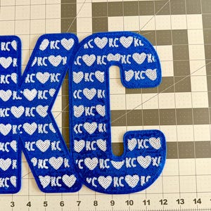 May include: Blue felt letters K and C with white sequins forming hearts and the letters KC. The letters are on a white and gray grid background.
