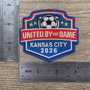 May include: A blue, red, and white embroidered patch featuring a soccer ball, the text UNITED BY THE GAME, KANSAS CITY, and 2026. A metal ruler shows the patch is approximately 3 inches wide and 3 inches tall on a wood grain surface.