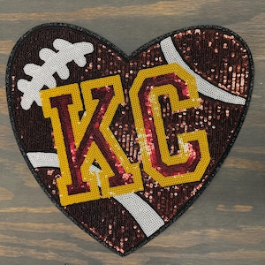 May include: A heart-shaped sequined patch in the style of a football. The patch is brown with white accents and features the letters "KC" in gold and maroon. The patch is likely for apparel or accessories.