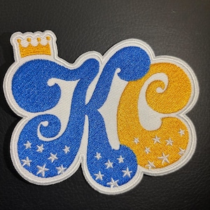 May include: A white, blue, and yellow embroidered patch with the letters "KC" in a stylized font. The letters are surrounded by white stars on a blue and yellow background. A gold crown sits above the letters.