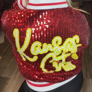 K&ouml;nnte beinhalten: Rote Paillettenjacke mit dem Schriftzug "Kansas City" in gelber und wei&szlig;er Schrift auf dem R&uuml;cken. Die Jacke hat einen wei&szlig;en Kragen mit roten und wei&szlig;en Streifen.