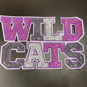 May include: A decorative patch with the word "WILDCATS" in large, block letters. The letters are outlined in purple and filled with sparkling rhinestones in various colors, including pink, black, and silver. The patch is on a gray background.