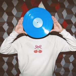 May include: A person is holding a blue vinyl record in front of their face. They are wearing a white sweatshirt with a pink cherry design.