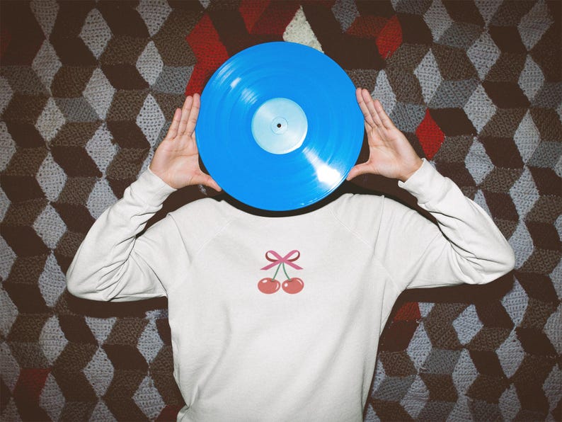 May include: A person is holding a blue vinyl record in front of their face. They are wearing a white sweatshirt with a pink cherry design.