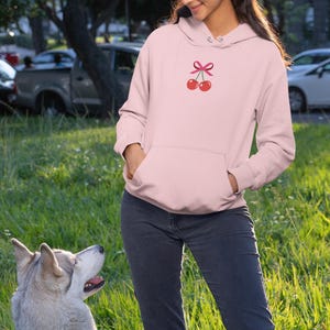 May include: A pink hoodie with a cherry design and a pink bow. The hoodie is worn by a person standing in a grassy area with a dog.