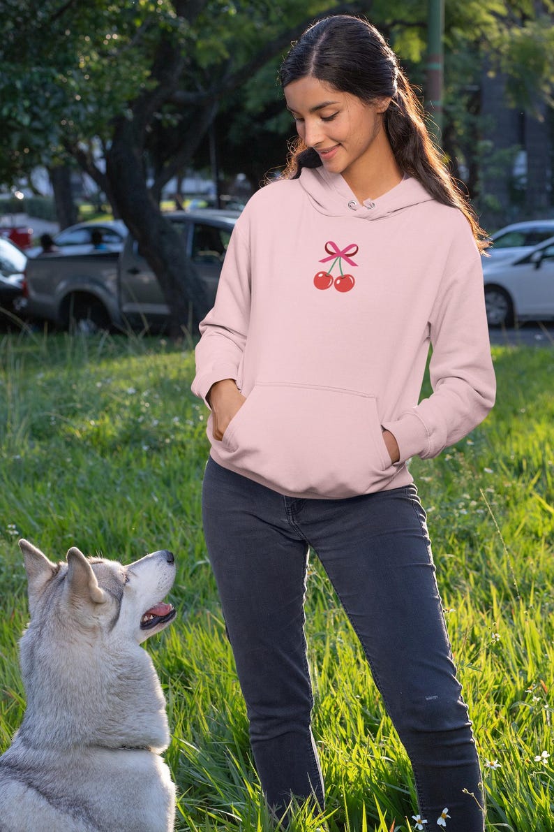 May include: A pink hoodie with a cherry design and a pink bow. The hoodie is worn by a person standing in a grassy area with a dog.