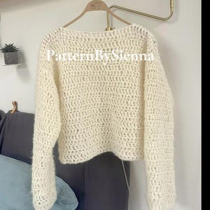 May include: A white crocheted sweater with a loose fit and long sleeves. The sweater has a simple, open-stitch design. The text "PatternBySienna" is visible on the front of the sweater.