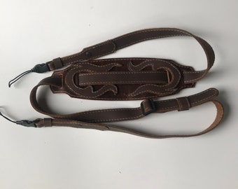 Könnte beinhalten: Ein braunes Lederkameraband mit einem dekorativen Design. Das Band hat zwei verstellbare Riemen und eine Schnellverschluss-Schnalle.