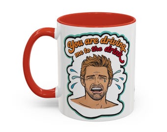 May include: A white ceramic mug with a red handle and interior rim. The front features a graphic of a crying person with brown hair and a beard, surrounded by a teal outline. The text reads, You are driving me to the drink, in orange and red.