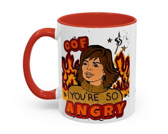 May include: A white ceramic mug with a red handle and interior rim. The front features a graphic illustration of a person with brown hair wearing a mustard yellow sweater, surrounded by orange flames, with the text OOF YOU'RE SO ANGRY.