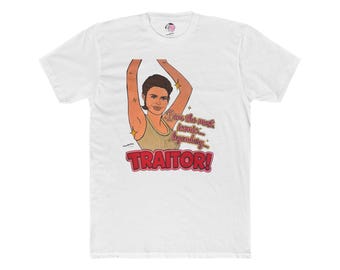 May include: A white short-sleeved t-shirt featuring a graphic illustration of a person with arms raised. The text reads, "I am the most iconic... legendary... TRAITOR!" in pink and red lettering with small yellow star accents surrounding the figure.