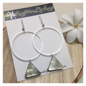 Puede incluir: Pendientes de aro de plata con un colgante de concha iridiscente en forma de triángulo. Los pendientes se muestran en una tarjeta blanca con el texto "Kreations By Kawai" y una pequeña flor blanca.