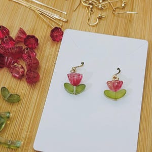 May include: A pair of handmade earrings featuring a tulip design. The earrings have a gold-colored hook and are composed of pink and green glass beads. Additional pink and green beads are scattered around the earrings on a wooden surface.