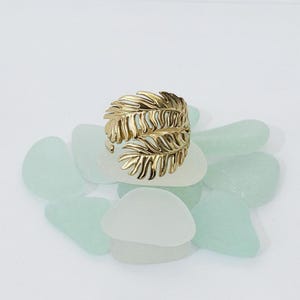 May include: A gold-toned ring featuring a wrap-around design shaped like two overlapping palm leaves with detailed veins. The metallic surface has a polished finish, and the band is open at the back, showcasing a botanical aesthetic.