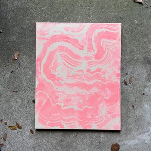 Puede incluir: Un lienzo rectangular con un diseño abstracto rosa y blanco. La pintura rosa se arremolina por el lienzo, creando un efecto marmolado. La obra de arte se exhibe sobre una superficie de hormigón gris.