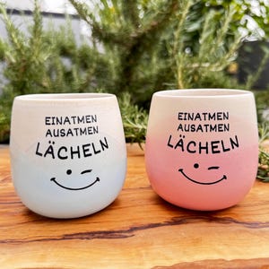 Könnte beinhalten: Zwei Keramikbecher mit abgerundeter Form. Ein Becher hat eine hellblaue Basis, der andere eine rosafarbene Basis. Beide Becher haben einen cremefarbenen oberen Teil und den schwarzen Text "EINATMEN AUSATMEN LÄCHELN" über einem zwinkernden Smiley.