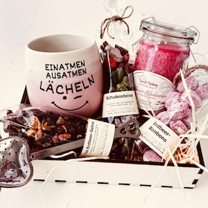 Könnte beinhalten: Ein Geschenkset in einer Holzkiste mit einer rosa Tasse mit der Aufschrift "EINATMEN AUSATMEN LÄCHELN" und einem Smiley, einem herzförmigen Tee-Ei aus Metall, losem Tee, Schokosteinen, einer rosa Kerze im Glas und Erdbeerbonbons.