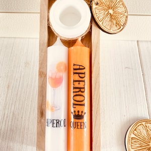 Op de afbeelding: Twee kaarsen in een houten doos. E&eacute;n kaars is wit met een illustratie van een Aperol Spritz cocktail. De andere kaars is oranje met het woord "APEROL" en een kroon erop gedrukt. De doos bevat ook een kleine houten sinaasappelschijf.