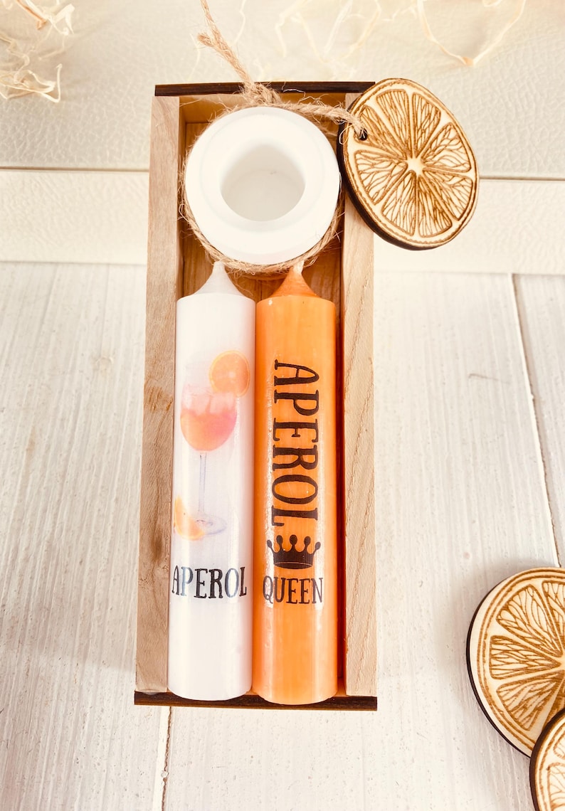 Op de afbeelding: Twee kaarsen in een houten doos. E&eacute;n kaars is wit met een illustratie van een Aperol Spritz cocktail. De andere kaars is oranje met het woord "APEROL" en een kroon erop gedrukt. De doos bevat ook een kleine houten sinaasappelschijf.