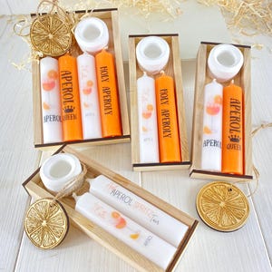 Op de afbeelding: Drie houten dozen, elk met twee kaarsen met oranje en witte ontwerpen. De kaarsen hebben de tekst "Aperol Queen", "Holy Aperol" en "Aperol Spritz" erop gedrukt. Elke doos bevat ook een klein houten sinaasappelschijfje.