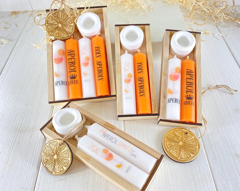 Op de afbeelding: Drie houten dozen, elk met twee kaarsen met oranje en witte ontwerpen. De kaarsen hebben de tekst "Aperol Queen", "Holy Aperol" en "Aperol Spritz" erop gedrukt. Elke doos bevat ook een klein houten sinaasappelschijfje.