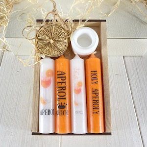 Op de afbeelding: Een houten doos met vier oranje kaarsen, elk met een ander Aperol Spritz ontwerp. De kaarsen zijn gelabeld met "Aperol Queen", "Eat, Drink, Aperol" en "Holy Aperol", en &eacute;&eacute;n kaars heeft een simpel Aperol logo. De doos bevat ook een houten sinaasappelschijf ornament.