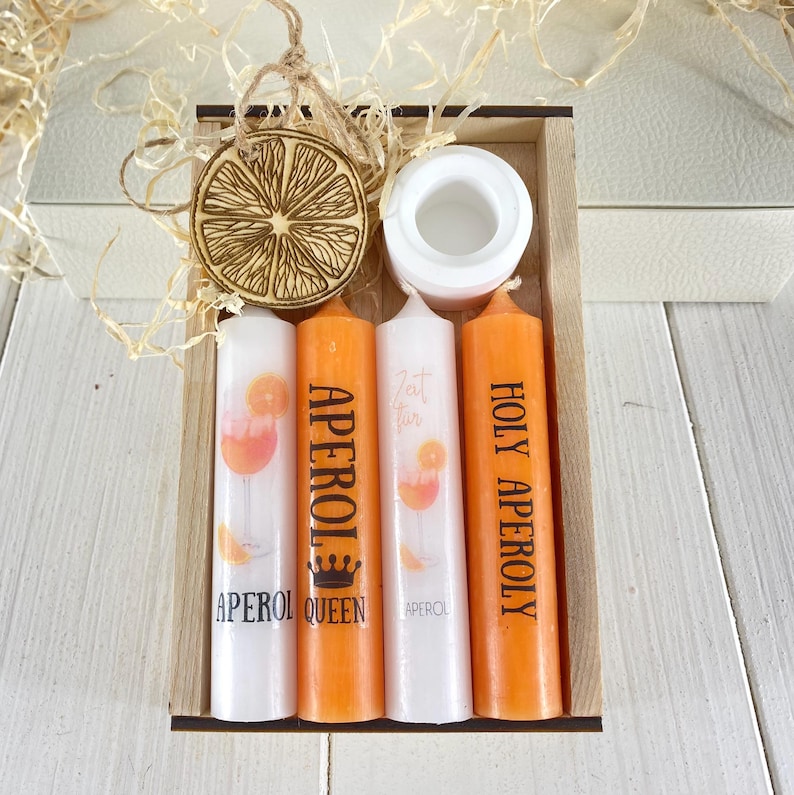 Op de afbeelding: Een houten doos met vier oranje kaarsen, elk met een ander Aperol Spritz ontwerp. De kaarsen zijn gelabeld met "Aperol Queen", "Eat, Drink, Aperol" en "Holy Aperol", en &eacute;&eacute;n kaars heeft een simpel Aperol logo. De doos bevat ook een houten sinaasappelschijf ornament.