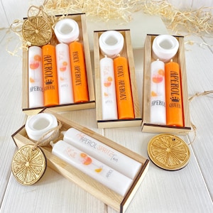 Op de afbeelding: Set van vier houten dozen met kaarsen erin. Elke doos bevat twee kaarsen, een witte en een oranje. De kaarsen zijn versierd met een glas sinaasappelsap en de tekst "Aperol" of "Holy Aperoly". De dozen zijn versierd met een houten sinaasappelschijf.
