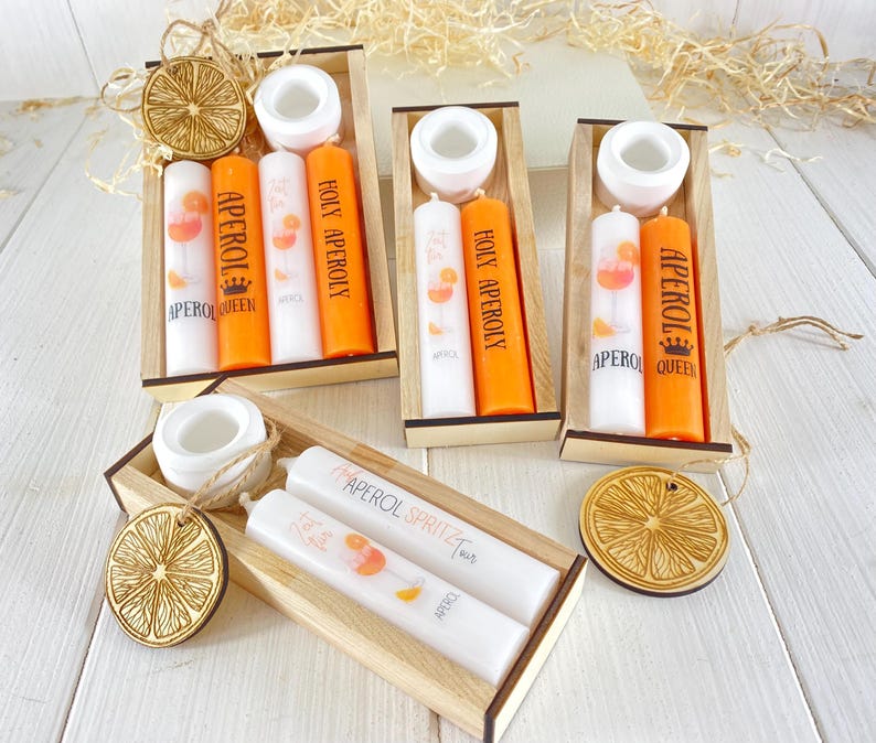 Op de afbeelding: Set van vier houten dozen met kaarsen erin. Elke doos bevat twee kaarsen, een witte en een oranje. De kaarsen zijn versierd met een glas sinaasappelsap en de tekst "Aperol" of "Holy Aperoly". De dozen zijn versierd met een houten sinaasappelschijf.