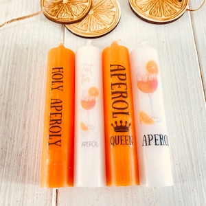 Op de afbeelding: Vier oranje en witte kaarsen met de tekst "Holy Aperoly", "Zeit f&uuml;r", "Aperol Queen" en "Aperol" erop gedrukt. De kaarsen zijn versierd met een afbeelding van een Aperol Spritz cocktail.