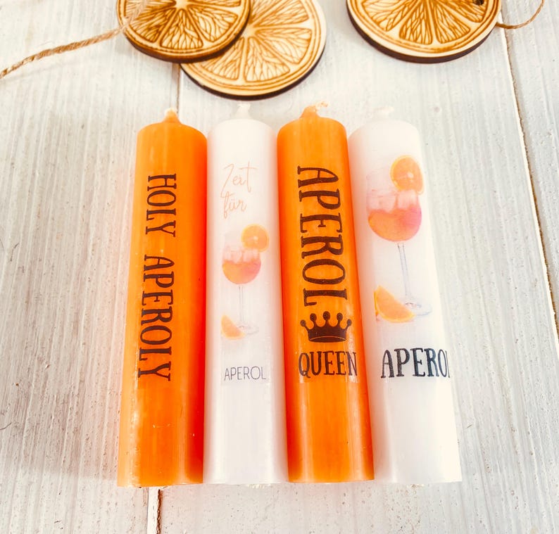 Op de afbeelding: Vier oranje en witte kaarsen met de tekst "Holy Aperoly", "Zeit f&uuml;r", "Aperol Queen" en "Aperol" erop gedrukt. De kaarsen zijn versierd met een afbeelding van een Aperol Spritz cocktail.