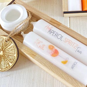 Op de afbeelding: Twee witte kaarsen in een houten doos. E&eacute;n kaars heeft de tekst "Aperol Spritz Tour" en de andere heeft de tekst "Eat, Drink, Fun".