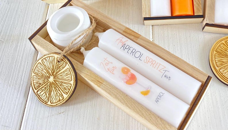 Op de afbeelding: Twee witte kaarsen in een houten doos. E&eacute;n kaars heeft de tekst "Aperol Spritz Tour" en de andere heeft de tekst "Eat, Drink, Fun".