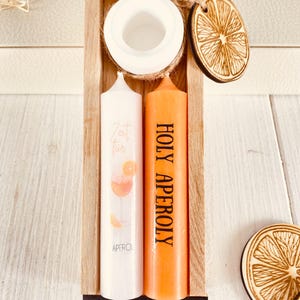 Op de afbeelding: Een houten doos met twee kaarsen erin. De kaarsen zijn wit en oranje. De witte kaars heeft een ontwerp van een Aperol Spritz cocktail. De oranje kaars heeft de tekst "HOLY APEROL" erop gedrukt. De doos bevat ook twee houten sinaasappelschijfjes.