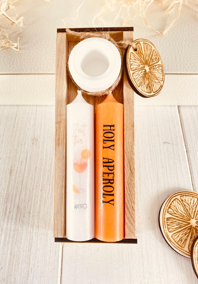 Op de afbeelding: Een houten doos met twee kaarsen erin. De kaarsen zijn wit en oranje. De witte kaars heeft een ontwerp van een Aperol Spritz cocktail. De oranje kaars heeft de tekst "HOLY APEROL" erop gedrukt. De doos bevat ook twee houten sinaasappelschijfjes.