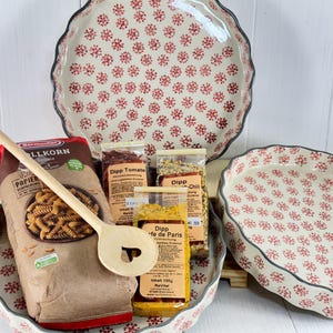 Könnte beinhalten: Drei weiße Keramik-Tortenplatten mit rotem Blumenmuster, eine Tüte Pasta, ein Holzlöffel und drei Gewürzpakete. Die Gewürzpakete sind mit "Dipp Tomate", "Dipp Chili" und "Dipp Cafe de Paris" beschriftet.