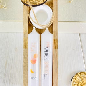 Op de afbeelding: Twee witte kaarsen met oranje en roze illustraties van een Aperol Spritz cocktail. De kaarsen bevinden zich in een houten doos met een houten deksel in de vorm van een sinaasappelschijf. De tekst op de kaarsen luidt "Zeit f&uuml;r Aperol Spritz".