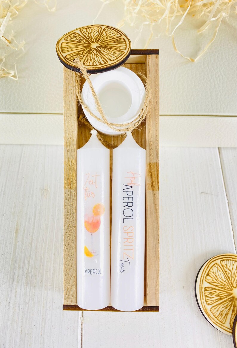 Op de afbeelding: Twee witte kaarsen met oranje en roze illustraties van een Aperol Spritz cocktail. De kaarsen bevinden zich in een houten doos met een houten deksel in de vorm van een sinaasappelschijf. De tekst op de kaarsen luidt "Zeit f&uuml;r Aperol Spritz".