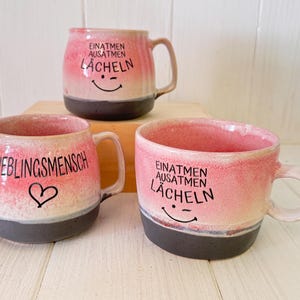 Könnte beinhalten: Drei Keramikbecher mit einem Farbverlauf in Rosa und Creme sowie einem dunkelgrauen Boden. Zwei Becher tragen den schwarzen Schriftzug "EINATMEN AUSATMEN LÄCHELN" mit einem lachenden Gesicht, einer trägt "LIEBLINGSMENSCH" mit einem Herzsymbol.