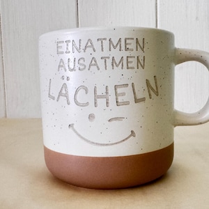 Könnte beinhalten: Ein Keramikbecher mit einem gesprenkelten cremefarbenen oberen Teil und einem terrakottafarbenen Boden. Auf der Vorderseite ist der Text "EINATMEN AUSATMEN LÄCHELN" eingraviert, ergänzt durch ein eingraviertes Smiley-Gesicht mit einem Zwinkern.