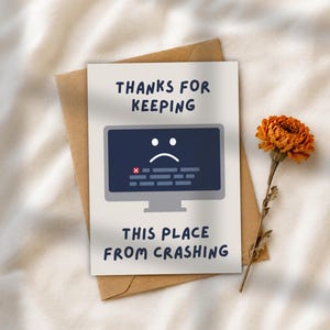May include: A white greeting card featuring a dark gray computer monitor graphic with a sad face icon and error text. The card displays the text THANKS FOR KEEPING THIS PLACE FROM CRASHING in dark blue letters, placed on a brown paper envelope.
