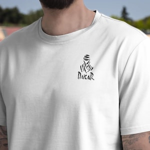 May include: White t-shirt with a black graphic of a figure in a head covering and the word "DAKAR" beneath. The design is located on the upper chest. The t-shirt is made of a soft material.