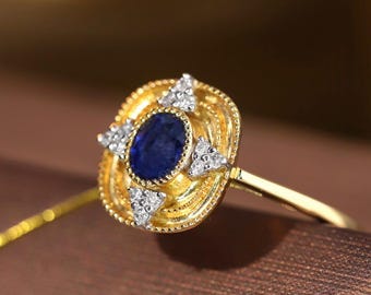 Op de afbeelding: Een gouden ring met een centrale ovaal geslepen blauwe saffier, omringd door een getextureerd, kralen gouden frame. Vier kleine driehoekige clusters van witte diamanten zijn op de windstreken rond de saffier geplaatst voor een gelaagd ontwerp.