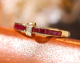 Può includere: Un anello in oro con una fila di rubini rosso scuro a taglio quadrato incastonati a canale. Il centro della fascia è accentuato da una barra verticale contenente tre piccoli diamanti chiari a taglio rettangolare.