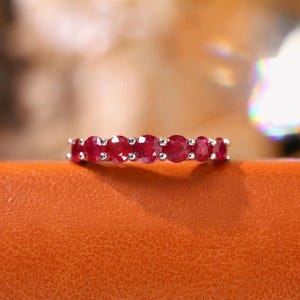 Pu&ograve; includere: Un anello color argento con una fila di sette pietre preziose rosse a taglio rotondo incastonate in una fascia metallica con griffe condivise. Le pietre sono uniformi per dimensioni e colore, creando una linea rossa continua sull'anello.