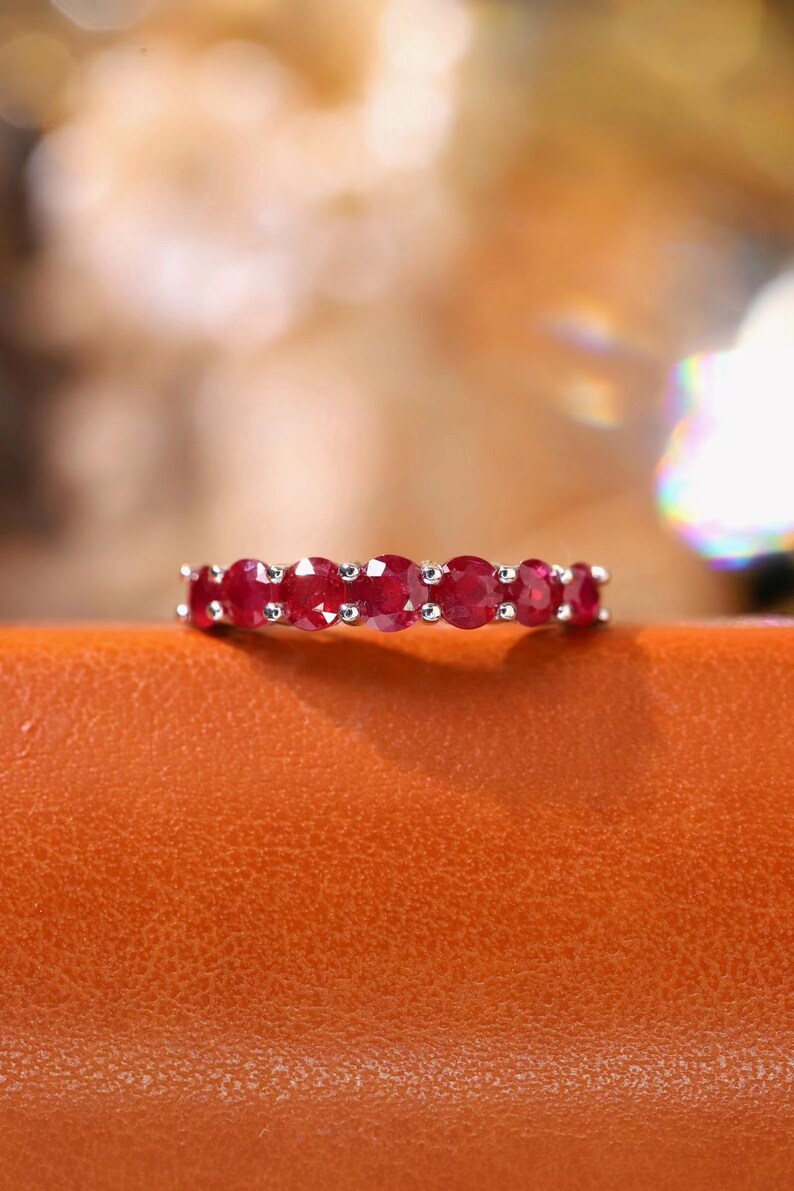 Pu&ograve; includere: Un anello color argento con una fila di sette pietre preziose rosse a taglio rotondo incastonate in una fascia metallica con griffe condivise. Le pietre sono uniformi per dimensioni e colore, creando una linea rossa continua sull'anello.