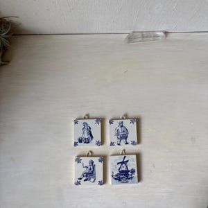 May include: Four square ceramic tiles featuring blue and white illustrations. Each tile depicts a different scene: a person with a basket, a person with a barrel organ, a person sitting in a chair, and a windmill. Each tile has a small hanging loop.