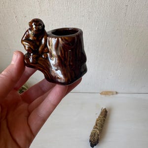 May include: A small, brown ceramic planter shaped like a tree stump with a monkey perched on the side. The planter has a glossy finish and is held in a person's hand. A bundle of sage and a crystal are in the background.