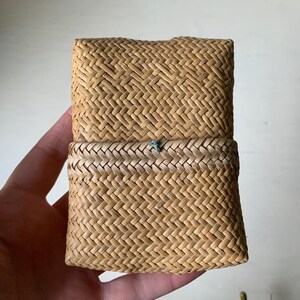 May include: A small, rectangular woven basket with a light brown colour and a woven pattern. The basket has a lid that is closed.